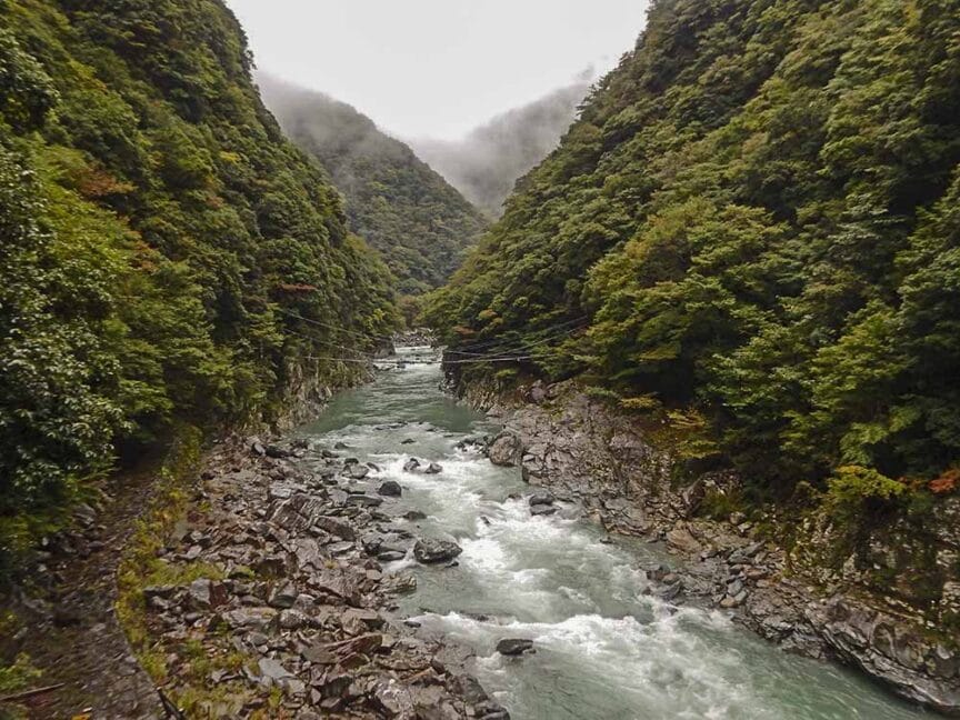 A river flows through a rocky gorge in Shikoku, surrounded by lush green vegetation and mist-covered hills. The water appears turbulent as it navigates between the steep, forested slopes, making it a perfect backdrop for hiking enthusiasts seeking adventure and history in nature.