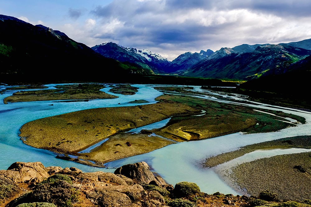 A winding river with multiple channels and islands flows through a mountainous landscape in Patagonia, leaving lasting impressions under a partly cloudy sky.