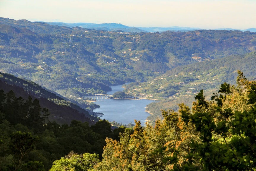 A river winds through a green valley surrounded by forested hills, with a bridge crossing the water in the distance under a clear sky, capturing the simple pleasures of rural Portugal.