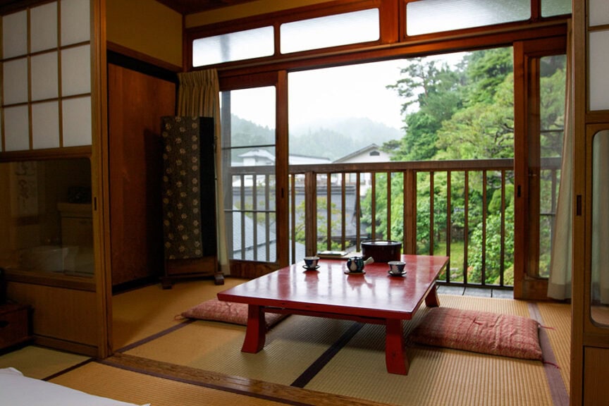 Traditional Japanese room with tatami mats, a low red table, and floor cushions. Sliding shoji screens open to a balcony with a view of greenery and buildings in the distance, evoking the serene ambiance of ancient Japanese culture akin to Koyasan.