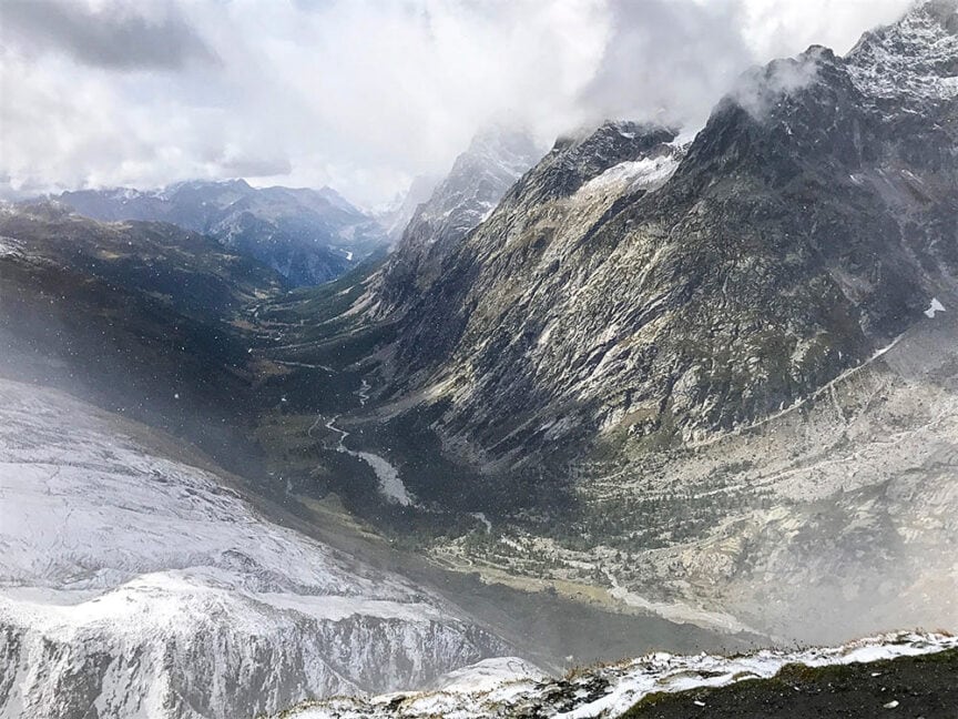 Snow-covered mountain range with a narrow river flowing through a green valley under a cloudy sky, perfect for hiking the iconic Tour du Mont Blanc.
