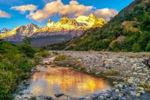 A serene mountain landscape in Patagonia with a rocky stream in the foreground. Snow-capped peaks are illuminated by a golden sunset, reflecting in the still water, surrounded by lush green forest. This picturesque scene is perfect for epic hikes and exploring national parks.