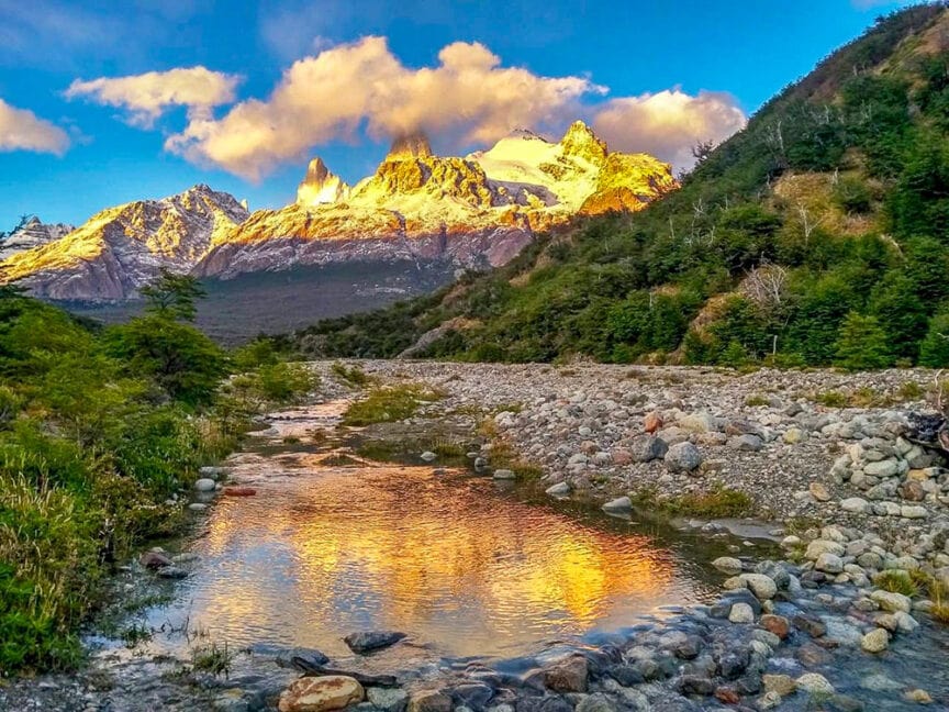A serene mountain landscape in Patagonia with a rocky stream in the foreground. Snow-capped peaks are illuminated by a golden sunset, reflecting in the still water, surrounded by lush green forest. This picturesque scene is perfect for epic hikes and exploring national parks.
