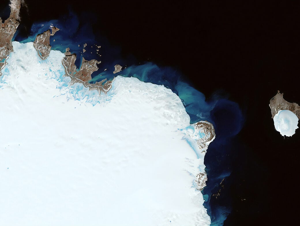 Stunning satellite image showing the northern coast and ice-covered terrain of Greenland with surrounding dark ocean waters and a smaller ice-covered island nearby on Earth.