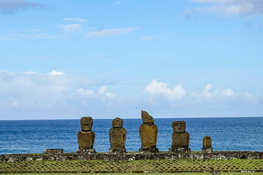Five large stone statues, known as moai, stand in a row on a grassy area near the ocean under a partly cloudy sky, evoking the island's ancient mysteries and tales of Tangata Manu.