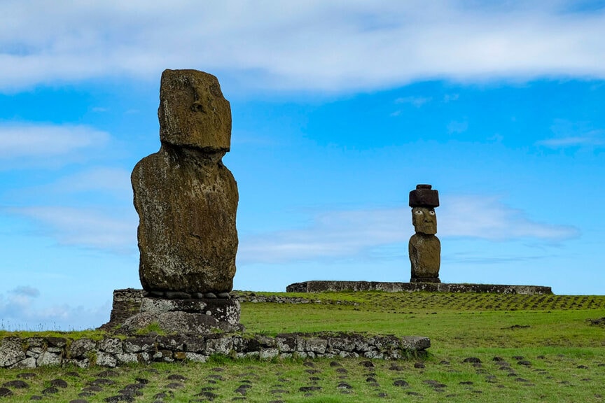 Two large stone Moai statues stand on platforms in a grassy field under a blue sky on Easter Island. One of the statues, embodying the island's timeless mysteries, is wearing a red stone topknot.