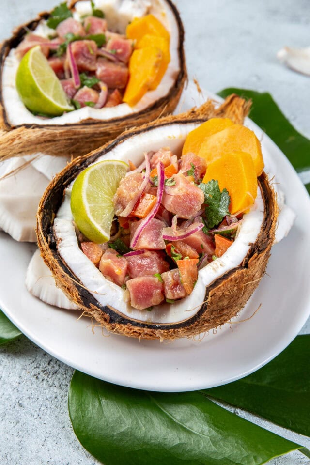 A dish of raw fish salad with sliced onions, herbs, lime, and yellow garnish served in a coconut shell on a white plate atop green leaves. Discover our delicious recipes to bring home flavors from around the world.