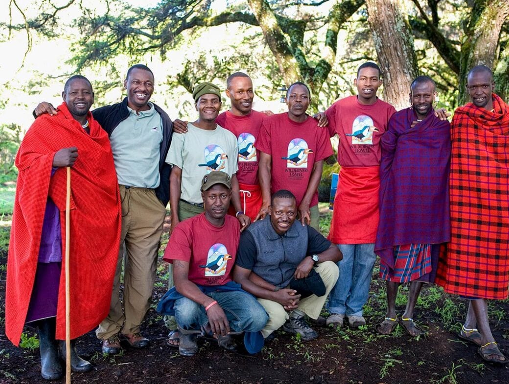 A group of men posing outdoors, some standing and some kneeling. Most are smiling, wearing red garments and T-shirts with a print. Trees and greenery form the backdrop, emphasizing the sense of community-based tourism in this vibrant setting.