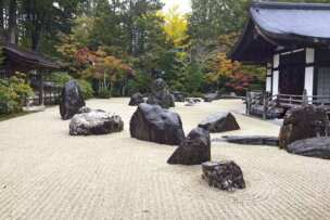 Zen garden with large rocks on raked gravel, surrounded by trees with autumn foliage, nestled on the historic island of Shikoku, and overlooked by traditional Japanese buildings.