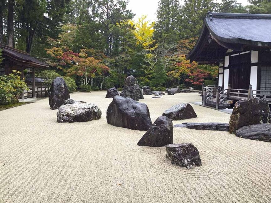 Zen garden with large rocks on raked gravel, surrounded by trees with autumn foliage, nestled on the historic island of Shikoku, and overlooked by traditional Japanese buildings.