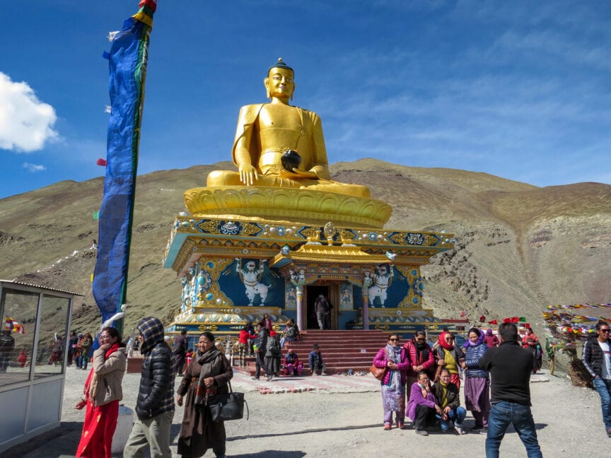 A large golden statue of Buddha is surrounded by people at a mountainous outdoor site in India. Colorful flags and fabric decorate the area, creating a vibrant atmosphere reminiscent of the majestic tigers and snow leopards that roam these regions.