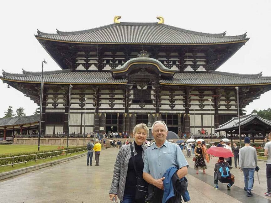 A couple stands in front of the historic Todai-ji Temple in Nara, Japan, on a cloudy day. Several other visitors can be seen around the temple grounds.