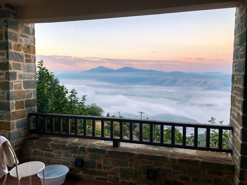 A stone balcony with a wooden railing overlooks a fog-covered landscape reminiscent of the isles near Greece, with distant mountains under a pastel sky. There's a white chair and small table on the balcony.