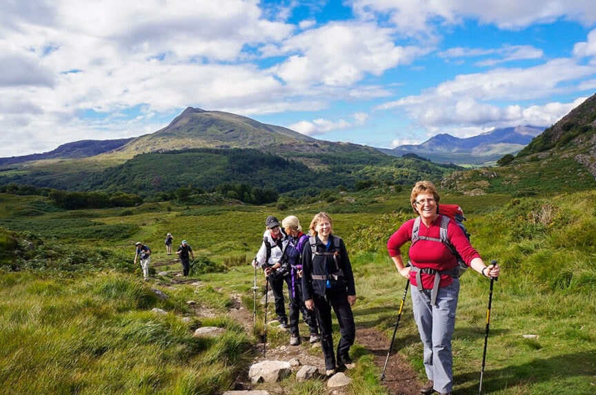 A group of travelers hiking on a trail through a verdant European landscape with mountains in the background under a partly cloudy sky.