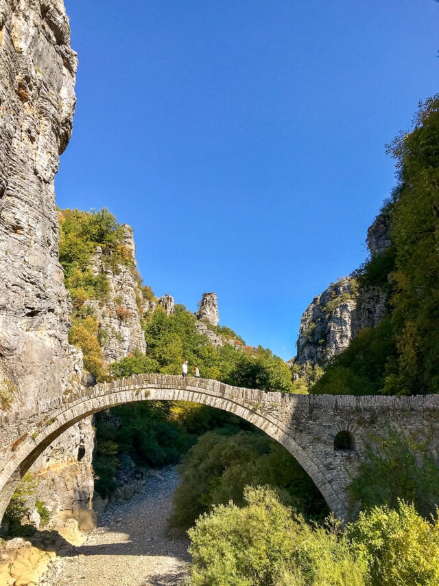 A stone arch bridge spans a dry riverbed between tall, rocky cliffs with green foliage under a clear blue sky, reminiscent of Greece's ancient architecture.