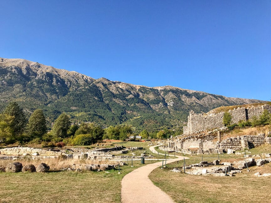A paved pathway leads through the ruins of an ancient site with stone structures, nestled in a mountainous landscape under a clear blue sky, reminiscent of Athens in Greece.