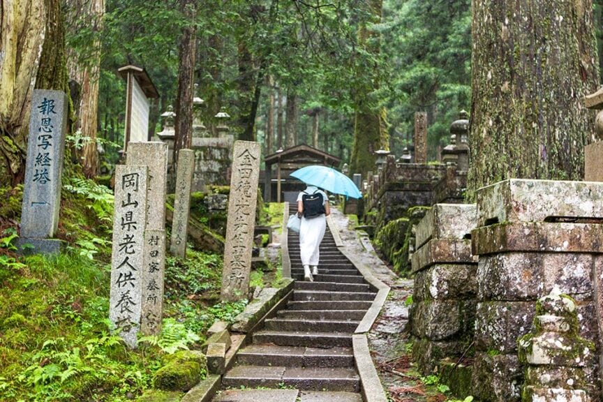 A person holding a blue umbrella walks up a stone staircase between moss-covered gravestones in a forested cemetery, exploring the serene beauty of Koyasan and its ancient Japanese culture.