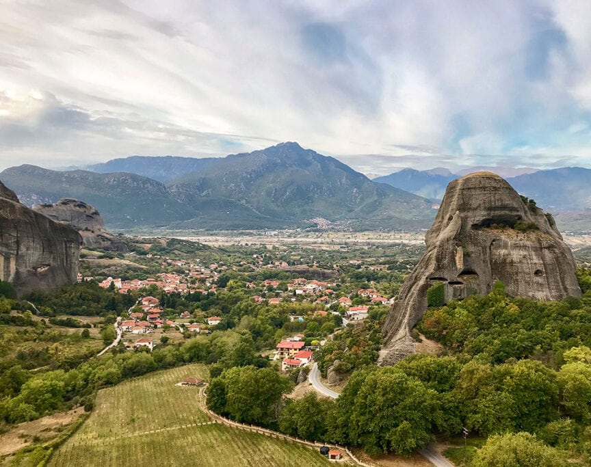 A scenic view of a countryside with lush green fields, scattered houses, large rock formations, and mountains in the background under a partly cloudy sky reminds one of exploring Greece, where similar serene landscapes can be found near Athens and throughout the Greek Isles.