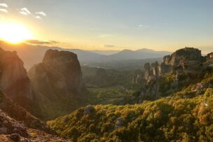 Sun setting over a mountainous landscape with rock formations, forests, and a valley in the distance. The scene evokes memories of the isles near Athens, where soft light and clear skies enhance the natural beauty.