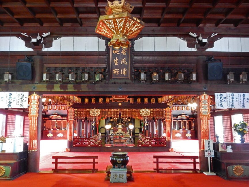 Interior of a traditional Japanese temple with ornate decorations, hanging lanterns, and wooden structures, reminiscent of the historical temples found on the Shikoku pilgrimage route. A red carpet covers the floor leading to an altar area adorned with various symbolic objects.