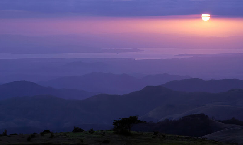 A sunset over a layered landscape of mountains and a body of water in the distance, with a tree visible in the foreground. The sky is tinted in shades of purple and orange.
