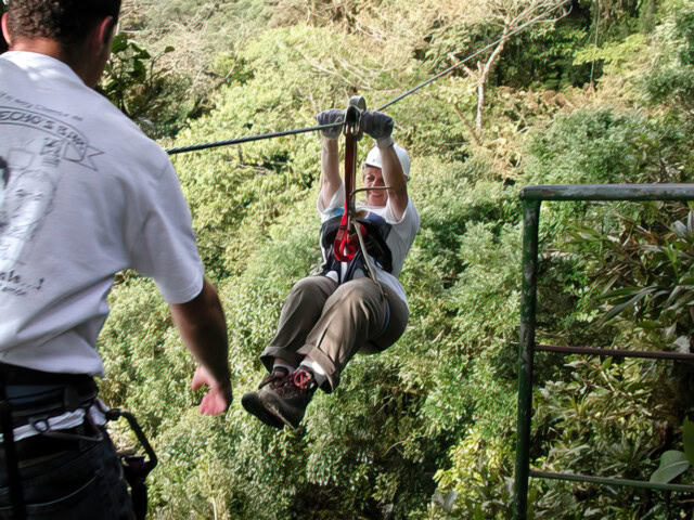 A person wearing safety gear is ziplining through a forested area, reminiscent of *Where the Wild Things Are*, while another person stands nearby assisting.