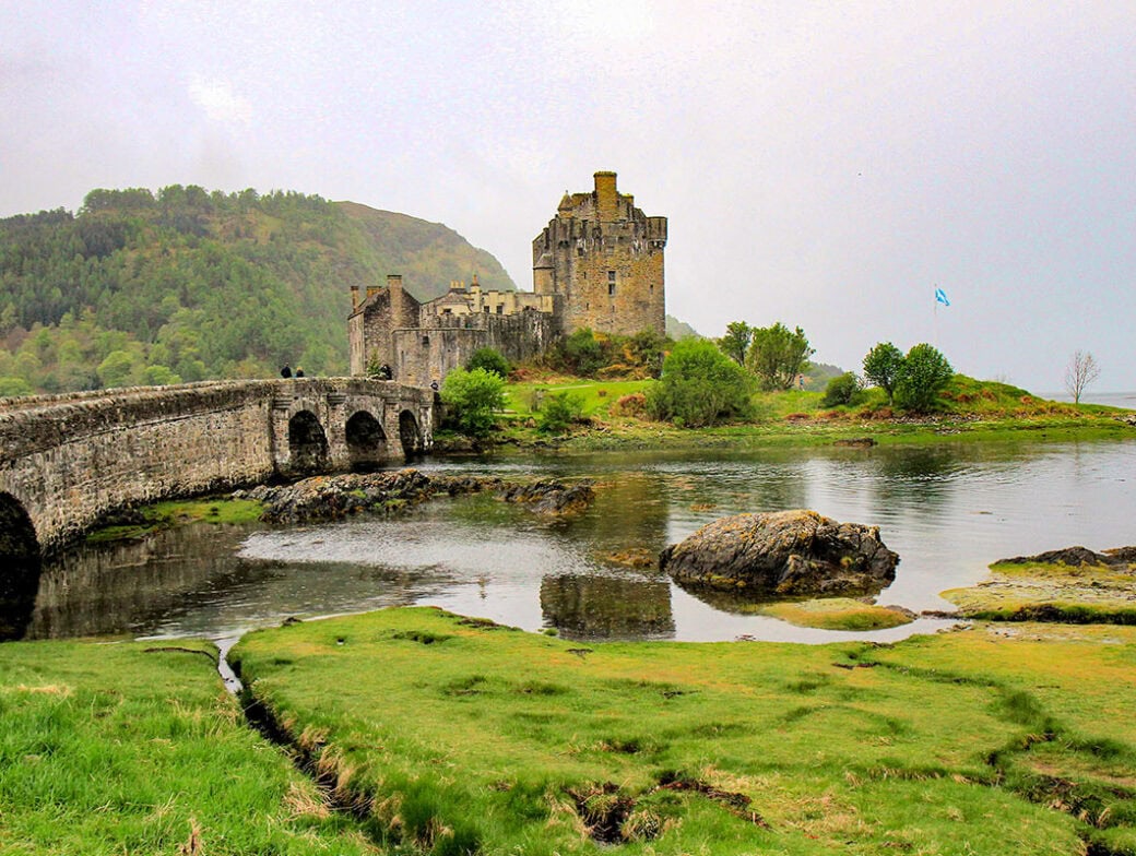 A stone castle on a small island is connected to the mainland by an arched stone bridge, with greenery and water in the foreground and wooded hills reminiscent of the Scottish Highlands in the background.