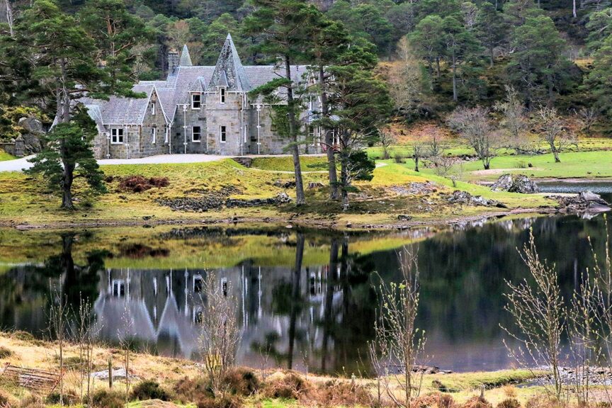 Stone mansion with steep gables and multiple chimneys set against a lush, forested hillside reminiscent of the Scottish Highlands. A tranquil pond in the foreground reflects the building and surrounding trees, creating a serene escape perfect for hiking enthusiasts.