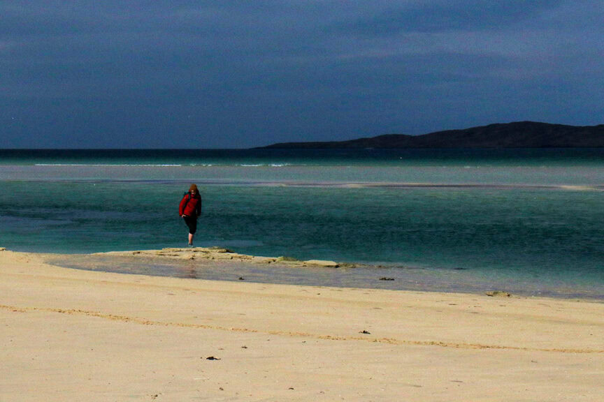 A person in a red jacket walks on a sandy beach with a calm sea and distant Scottish Highlands under a cloudy sky.