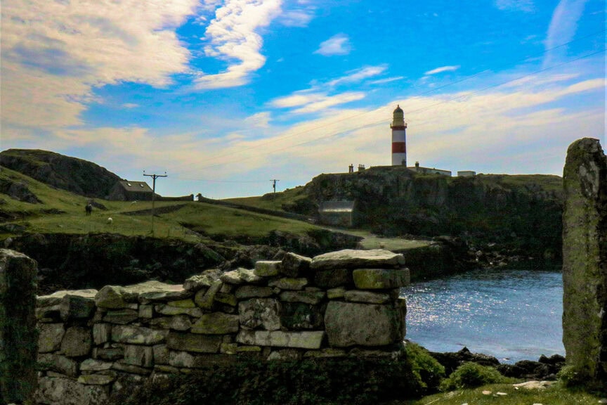 A scenic view of a coastal landscape in the Scottish Highlands featuring a stone fence in the foreground, a body of water, and a red-and-white striped lighthouse in the background under a partly cloudy sky—an ideal spot for hiking enthusiasts.