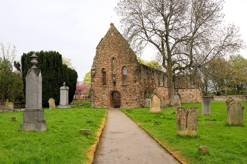 An old, stone church building stands at the end of a path surrounded by gravestones and greenery, with a leafless tree nearby. The scene evokes the rugged beauty of the Scottish Highlands, inviting you to imagine an adventurous hike through its storied past.