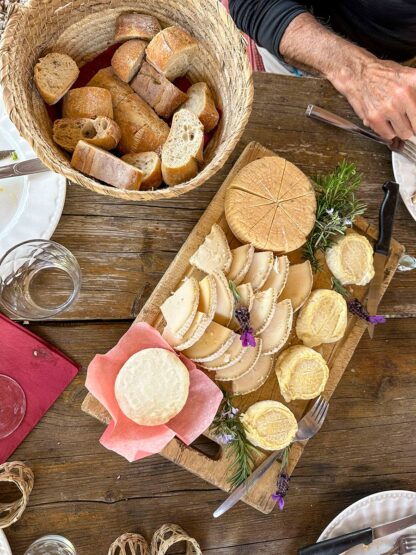 A wooden table set for a meal, reminiscent of adventures on the Islands of Corsica, features a basket of sliced bread and a cutting board with assorted cheeses and garnishes. Plates, glassware, and utensils are also present.