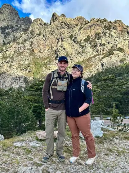 Two people stand close together, smiling, in a mountainous outdoor setting. They are dressed in casual trekking clothes and backpacks, with rocky cliffs and greenery in the background, capturing their Corsica adventures.