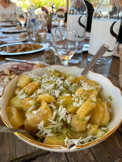 A bowl of sliced apples garnished with white flowers and herbs on a rustic table set with plates, glasses, and water bottles, evokes the charm of culinary adventures in Sardinia.