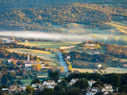 A scenic view of a rural countryside with houses, a winding road, green fields, dense forests, and a layer of mist in the distance evokes the adventures waiting in Corsica and Sardinia.