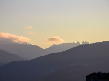 A view of mountain ranges at sunset with layers of peaks and a partially cloudy sky, seen from a vantage point including a shadowed building in the lower right corner, reminiscent of island adventures in Sardinia.