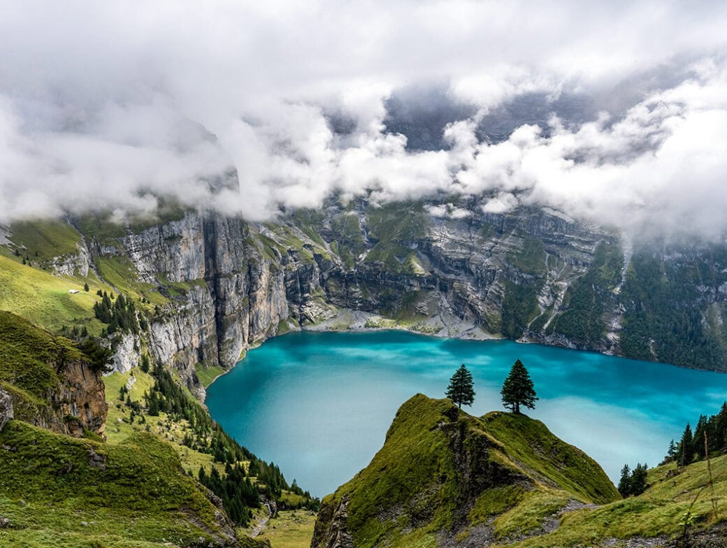 A scenic travel view of a turquoise lake surrounded by steep cliffs and green hills in Switzerland, with clouds partially covering the sky. Two trees stand prominently in the foreground on a grassy hill, capturing the essence of September's serene beauty.