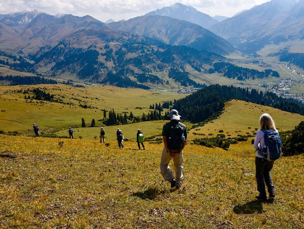 A group of hikers with backpacks descends a grassy hillside, overlooking a valley with forested areas and distant mountains under a partly cloudy sky—an invitation to explore Kyrgyzstan's breathtaking landscapes.