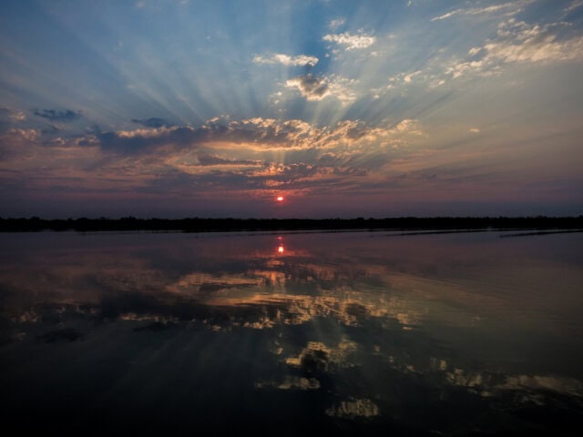 Sunset over a calm lake in Botswana with sun rays breaking through clouds, reflected in the water below, showcases the best of Botswana's natural beauty, making it a perfect travel blog feature.