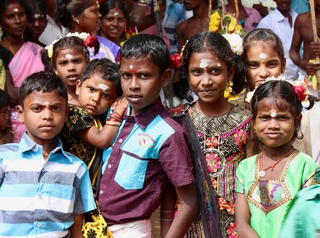 A group of children from South India, adorned with traditional attire and white forehead marks, poses for a portrait in a crowded setting.
