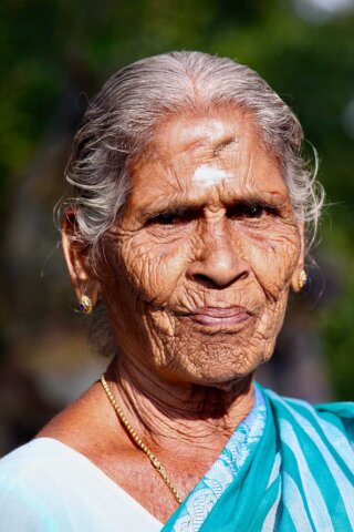 An elderly woman with gray hair and wrinkles, wearing a white and blue sari and gold jewelry, looks at the camera with a neutral expression in this South Indian portrait. The background is blurred with greenery.