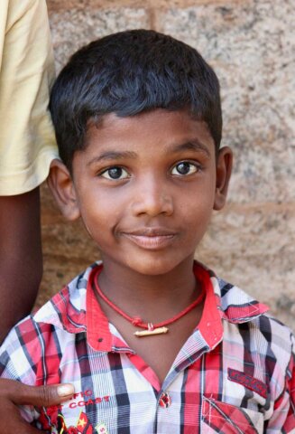 A young boy with short black hair, wearing a red and white plaid shirt and a red necklace, stands against a brick wall with part of another person visible behind him. The scene captures the essence of traditional South Indian portraits.