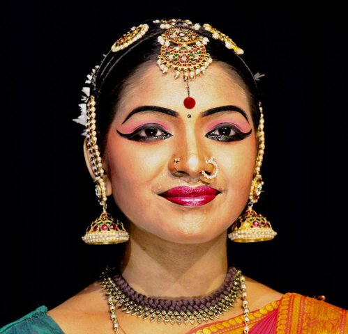 A captivating portrait of a person adorned in traditional South Indian dance attire with intricate jewelry, including a headpiece, nose ring, and large earrings. The elaborate makeup highlights prominent eye and lip details, all set against a striking black background.