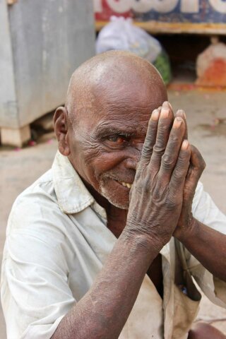An elderly man with a bald head and wearing a light-colored shirt holds his hands together near his face, smiling. This charming portrait captures him in an outdoor setting, reminiscent of the warm ambiance of South India.