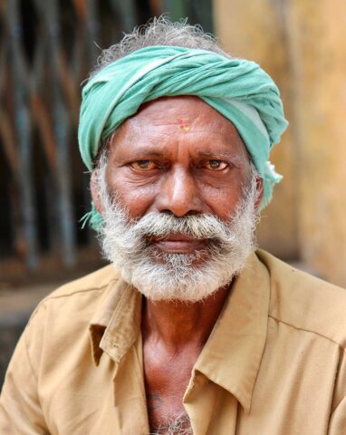 A man with a white beard and mustache wears a green headscarf and a beige shirt, looking directly at the camera. This striking portrait captures the essence of South India.