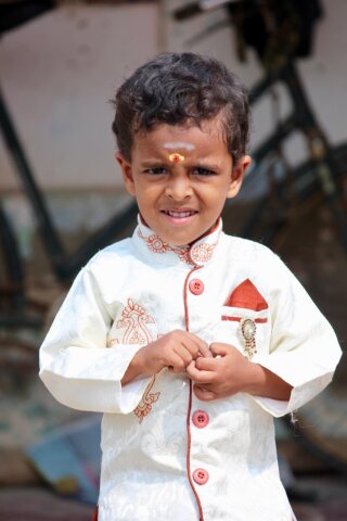 A young boy with curly hair, wearing a white traditional outfit with red buttons and embroidery, stands outdoors in this captivating South India portrait, a slight smile playing on his lips and a decorative mark adorning his forehead.