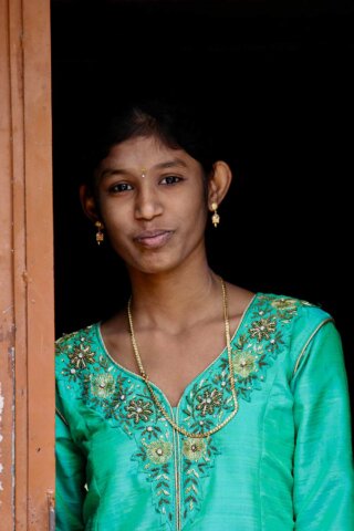 A young woman in a green embroidered top with gold jewelry stands in a doorway, looking directly at the camera against a dark background. This captivating portrait captures the essence of South India.