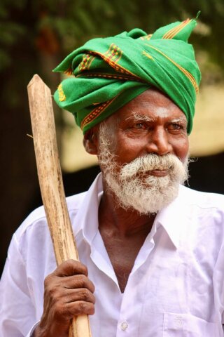 Elderly man with a white beard wearing a green turban and white shirt, holding a wooden stick, and standing outdoors. This captivating portrait captures the essence of South India.