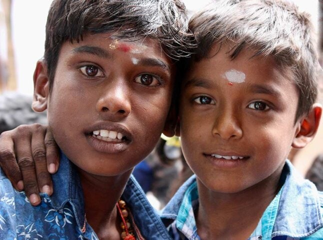Two boys, one with his arm around the other, smiling at the camera. Both have markings on their foreheads and are wearing blue shirts. This beautiful portrait captures the essence of South India through photography.