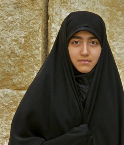 A person wearing a black hijab stands in front of a textured stone wall, embodying the beauty of Iran's culture as they look directly into the camera with warmth in their eyes.